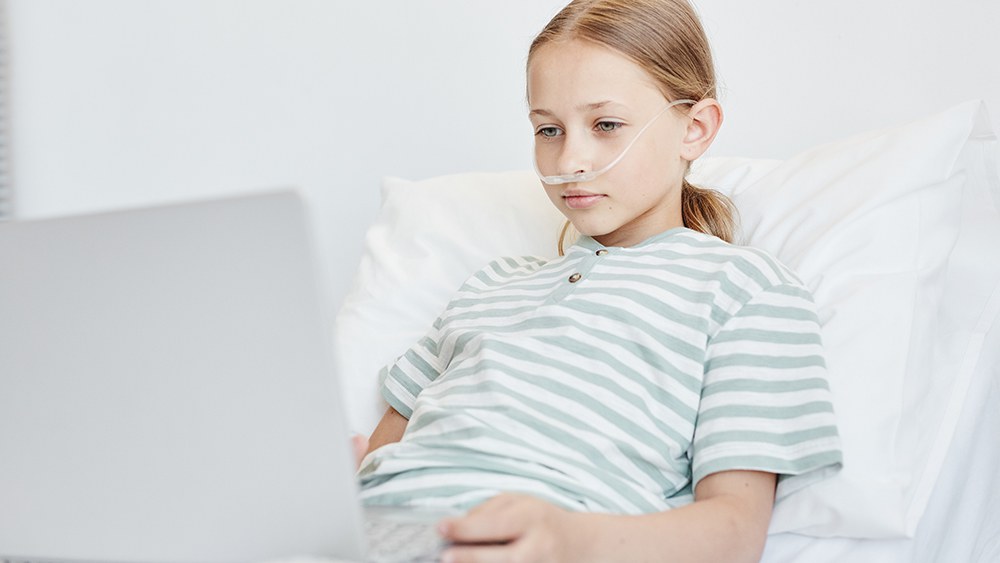 Krankes Mädchen liegt im Spitalbett und schaut in einen Laptop