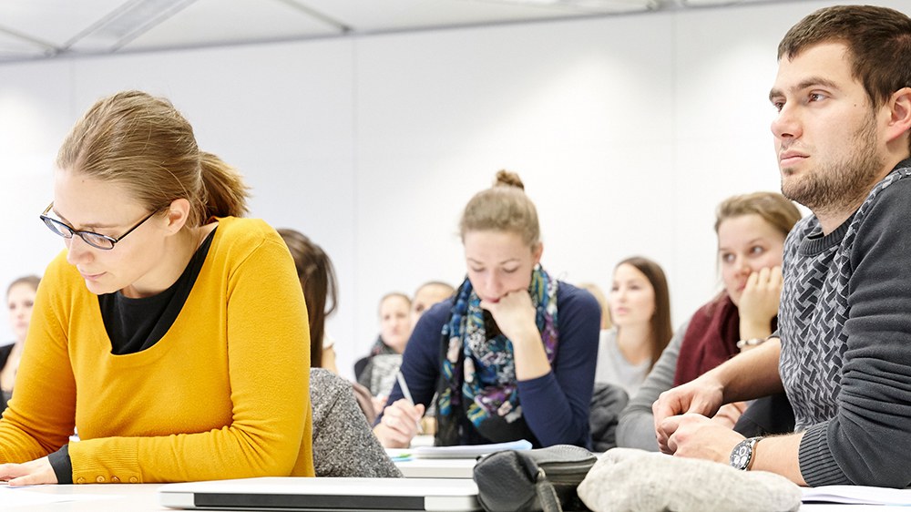 Studierende sitzen an Tischen, machen Notizen und hören aufmerksam zu