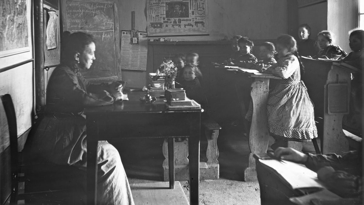 Blick auf eine Schulklasse in einer Tessiner Dorfschule um 1920; am Pult sitzt eine Lehrerin, etwa zehn Kinder sitzen in Bänken.
